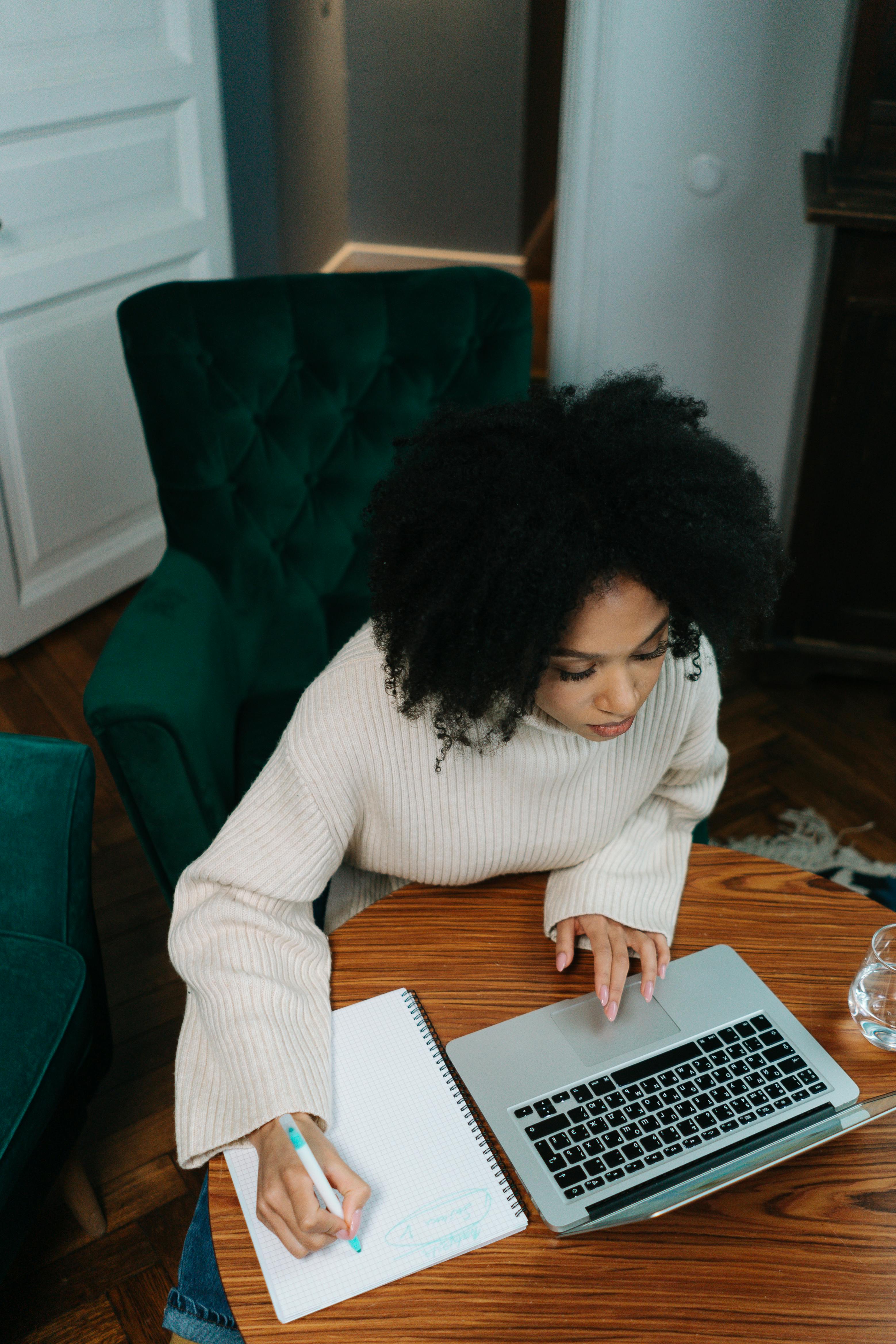 Woman sitting at desk, using computer and writing in notebook