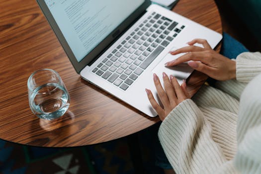 A person working on a laptop at a wooden table with a glass of water.