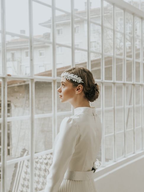 Pensive young female in white elegant dress and hairband standing near windows in room in daylight
