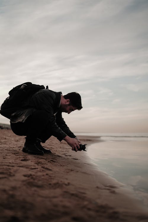 Side view full length of guy in black clothes and backpack sitting on sandy beach near ocean under cloudy gray sky in daylight