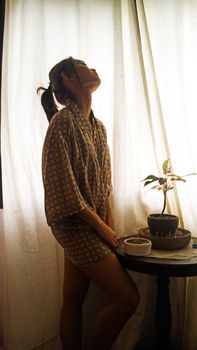 A woman in patterned pajamas stands by a sunlit window with a houseplant on a table.
