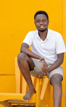 Smiling man in white polo shirt and khaki shorts sitting on a chair against a vibrant yellow background.