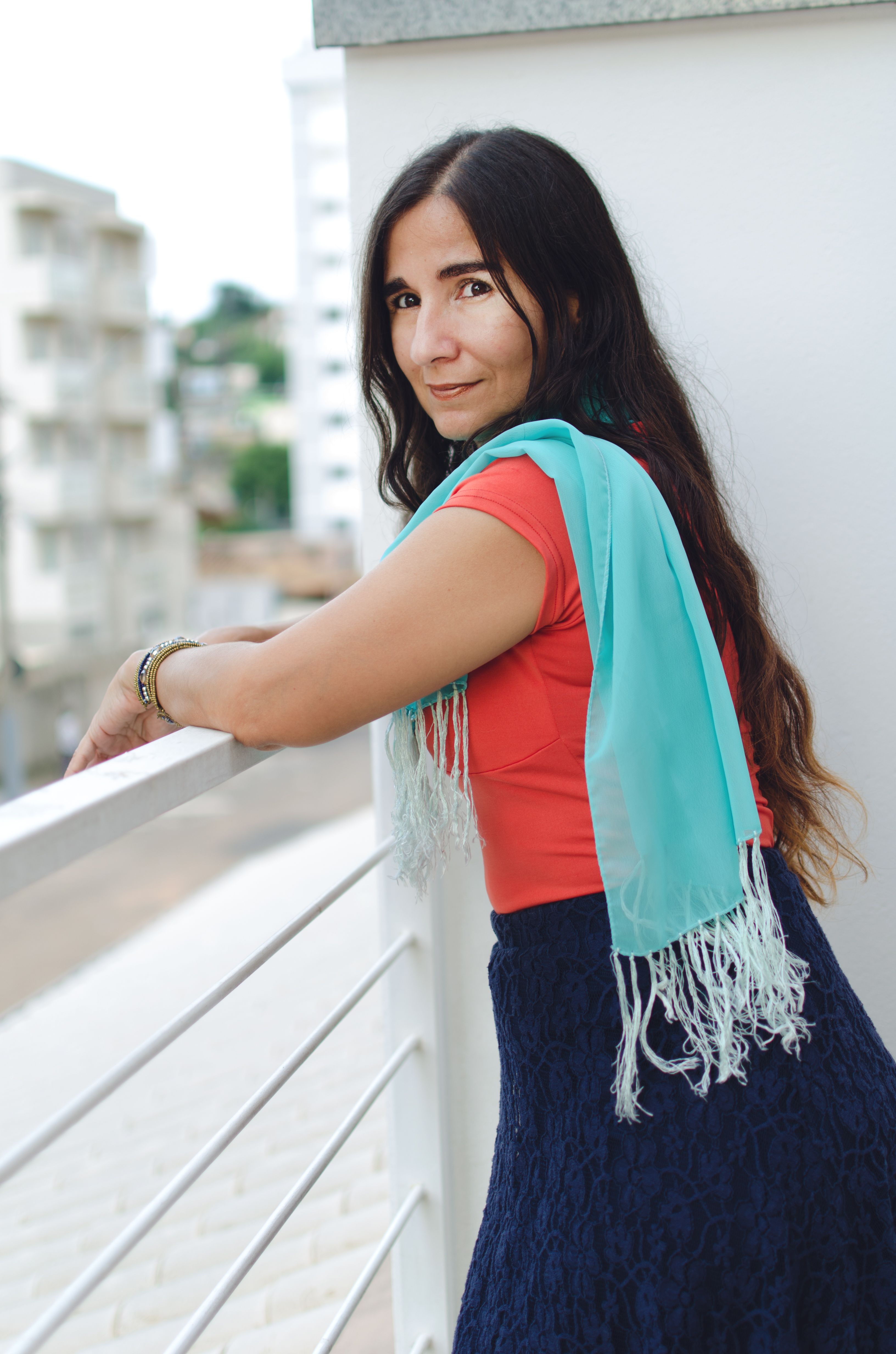 A woman stands on a balcony in Juiz de Fora, Brazil, smiling warmly with a scenic city view.