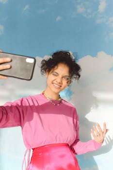 Smiling teenage girl in pink sweater taking a selfie with a cloudy sky backdrop, showcasing joyful and vibrant expression.
