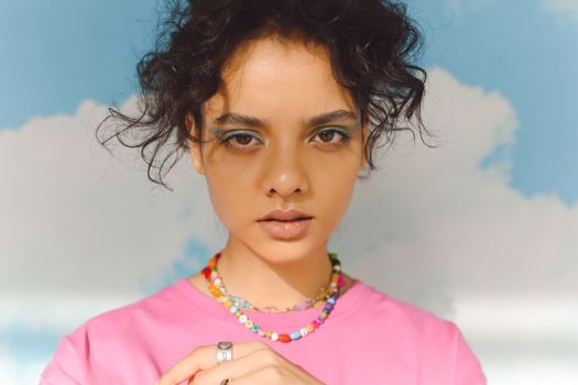 Close-up portrait of a woman with curly hair and colorful necklace against blue sky backdrop.