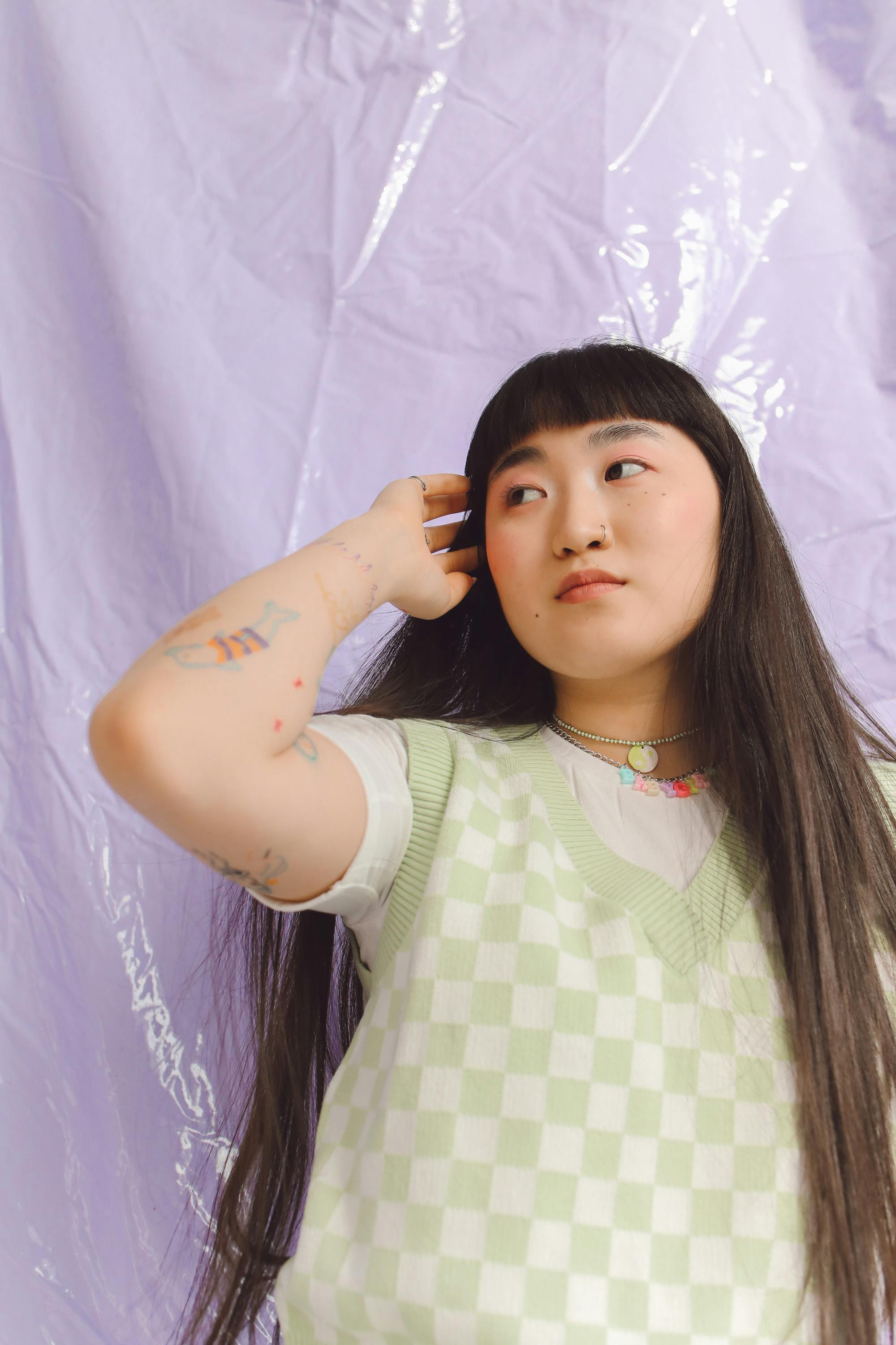 Asian woman posing against a lavender backdrop, wearing a green checkered vest.