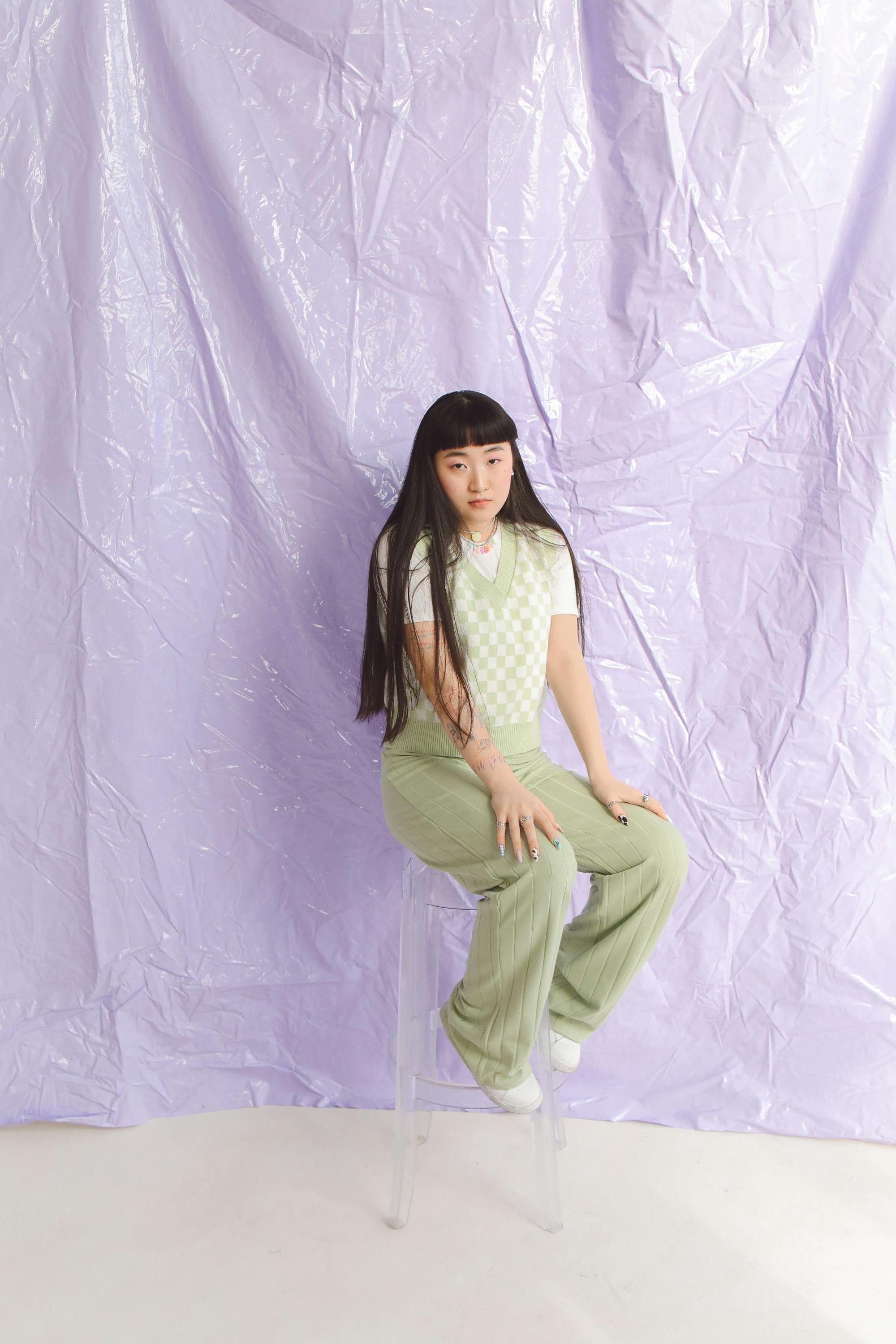 Young woman seated on clear chair against pastel backdrop, showcasing minimalist fashion.