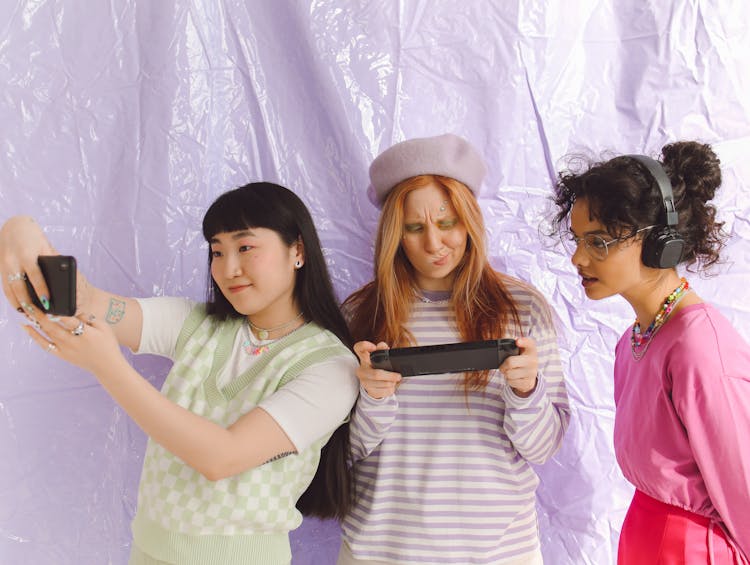 Three Girls With A Smart Phone, A Handheld Game Console And Headphones