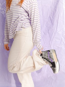 A person in a striped long-sleeve shirt and white pants showcases trendy zebra-print sneakers against a purple backdrop.