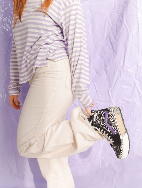 A person in a striped long-sleeve shirt and white pants showcases trendy zebra-print sneakers against a purple backdrop.