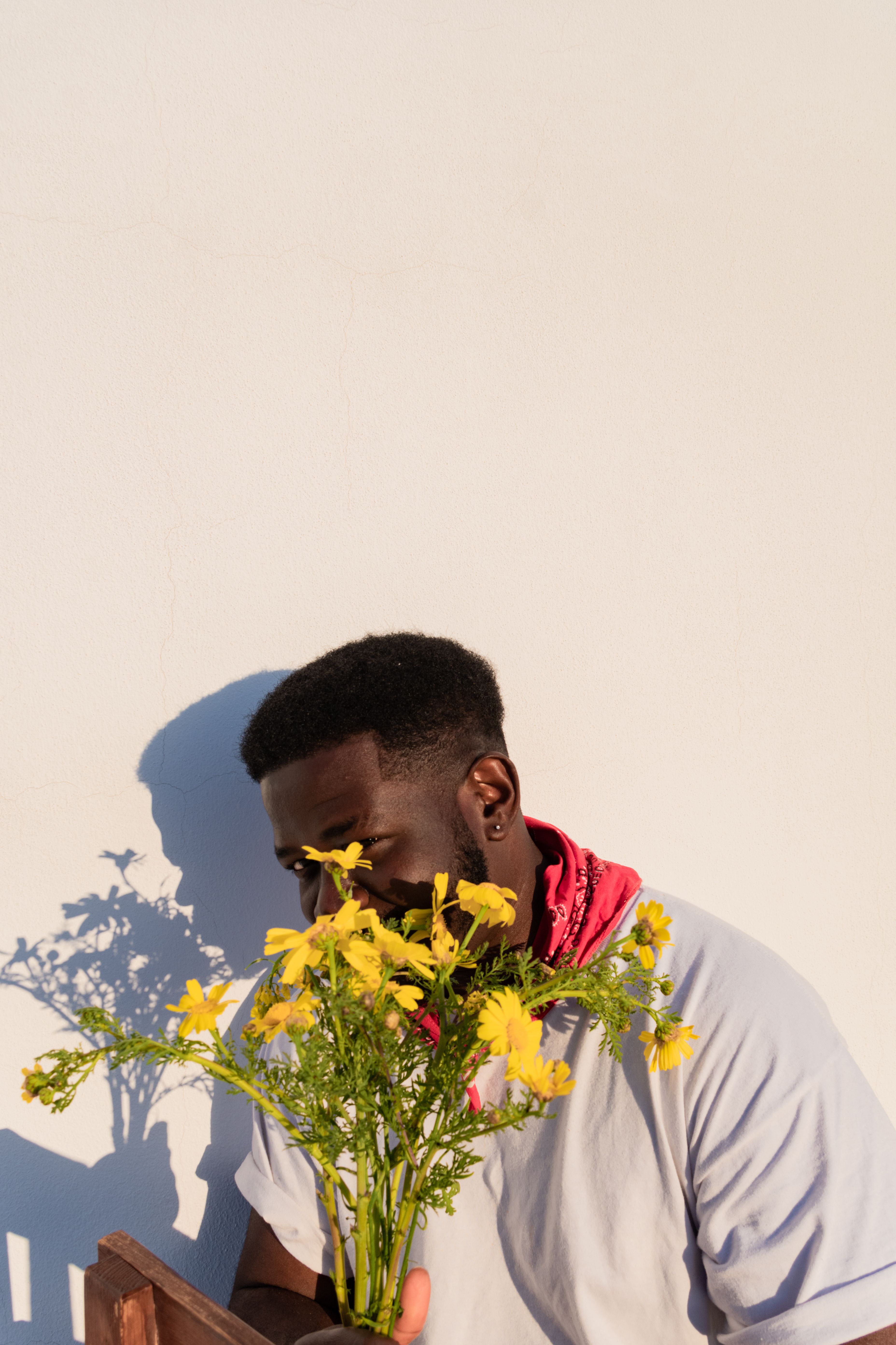 African man partially hiding behind yellow flowers, sunlit wall.