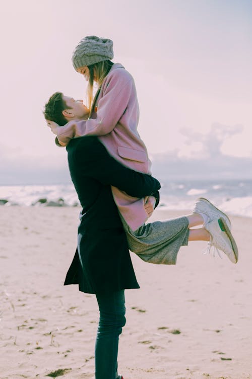 Man in Gray Coat Carrying Woman Wearing Pink Coat in Beach Near Shoreline and Body of Water