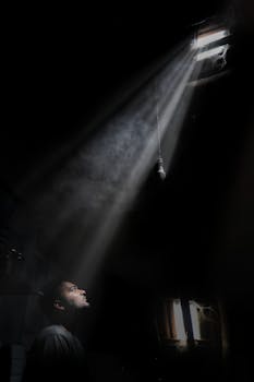 A contemplative man gazes at a sunbeam piercing through a dark room's ceiling.