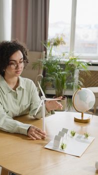 Woman with curly hair examines a model wind turbine for a school project indoors.