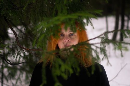 Free Pensive adult woman in black coat standing near green fir twigs in snowy forest and looking at camera in winter day Stock Photo