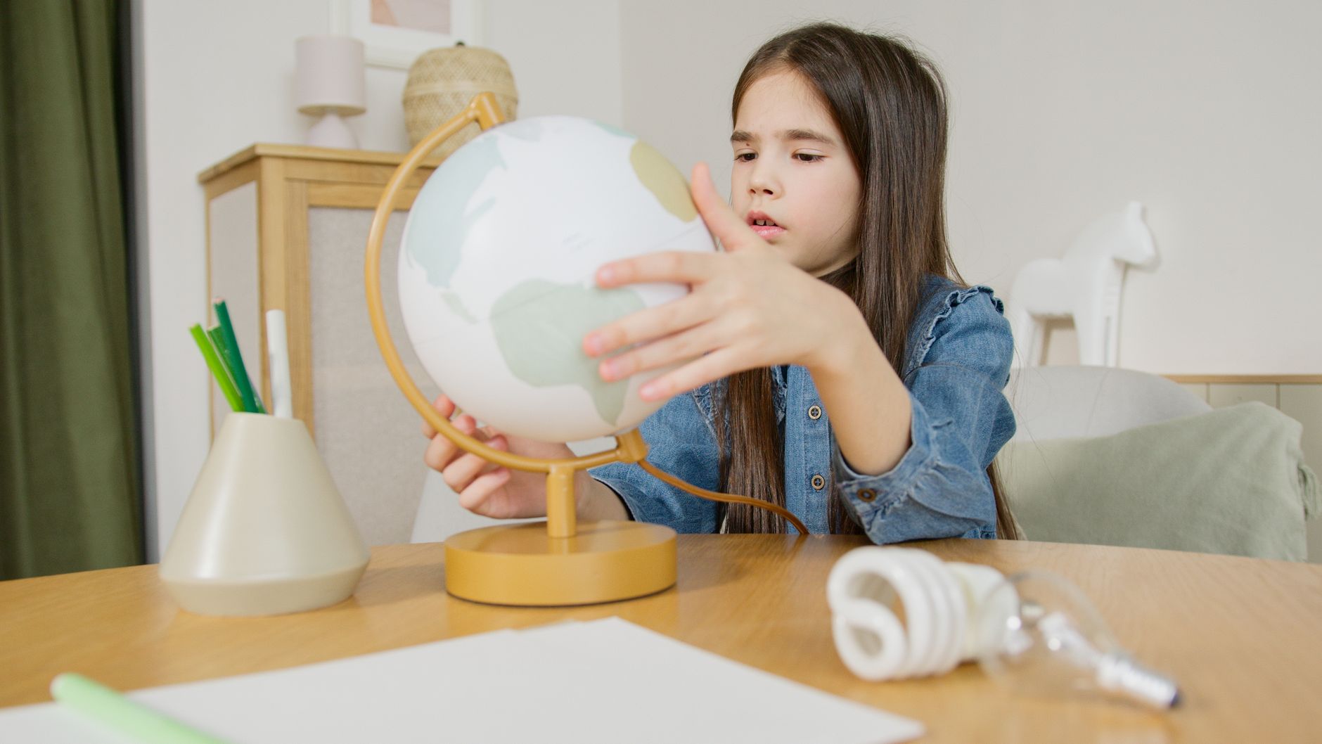 A young girl in a denim shirt examines a globe at a desk indoors, symbolizing learning and exploration.