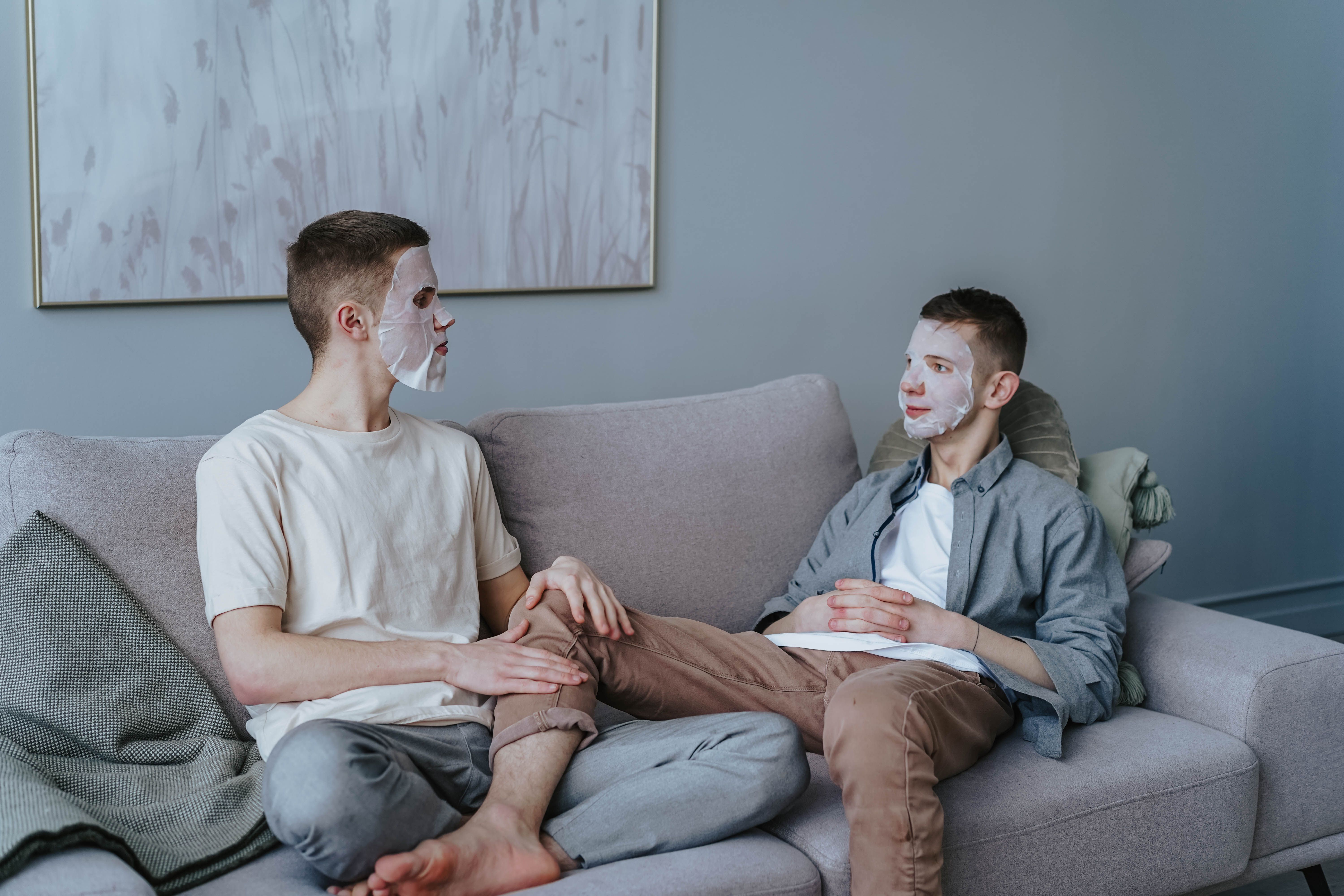 Two men sitting on a couch with face masks, sharing a relaxed skincare moment indoors.