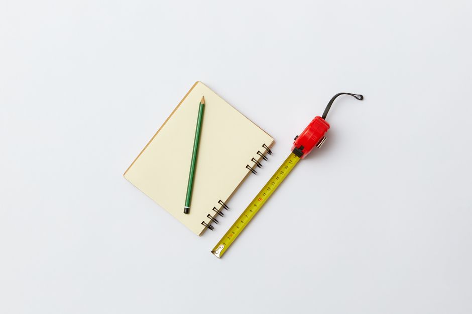 Simple flat lay of a notebook, green pencil, and red tape measure on a white background.