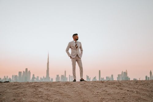Free A man in a suit standing in a desert with the Dubai skyline at sunset, showcasing a modern urban backdrop. Stock Photo