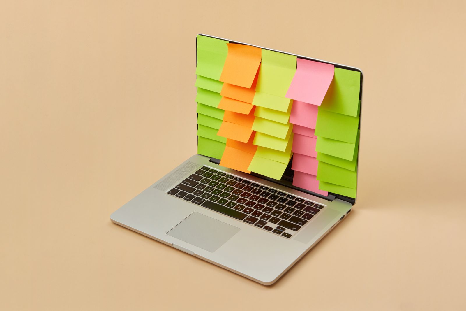An open silver laptop on a plain beige background, its entire screen covered with rows of orange, green and pink sticky notes overlapping each other