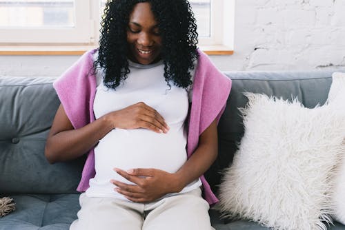 Cheerful pregnant African American female touching tummy gently while sitting on comfortable sofa with pillows in light room at home