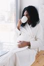Calm pregnant African American female in bathrobe drinking beverage from cup while looking out window in light room at home