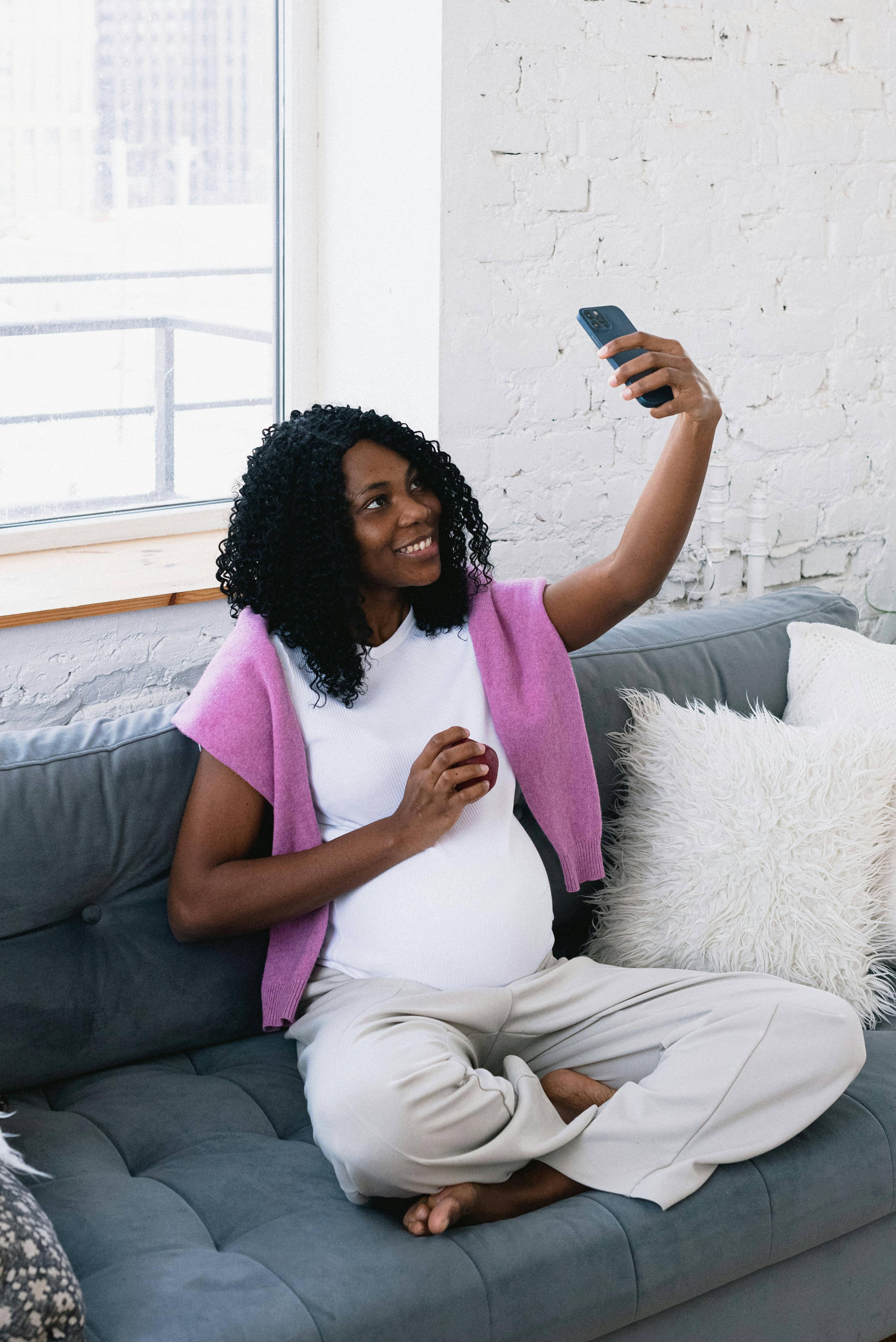 cheerful pregnant black woman taking selfie on couch