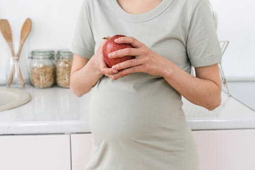 Free Unrecognizable pregnant female with ripe red apple for healthy diet in hands standing near counter in light kitchen at home Stock Photo