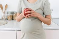 Unrecognizable pregnant female with ripe red apple for healthy diet in hands standing near counter in light kitchen at home