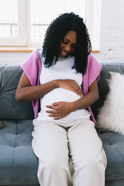 Joyful pregnant African American female with black hair touching tummy gently while sitting on sofa in light living room at home