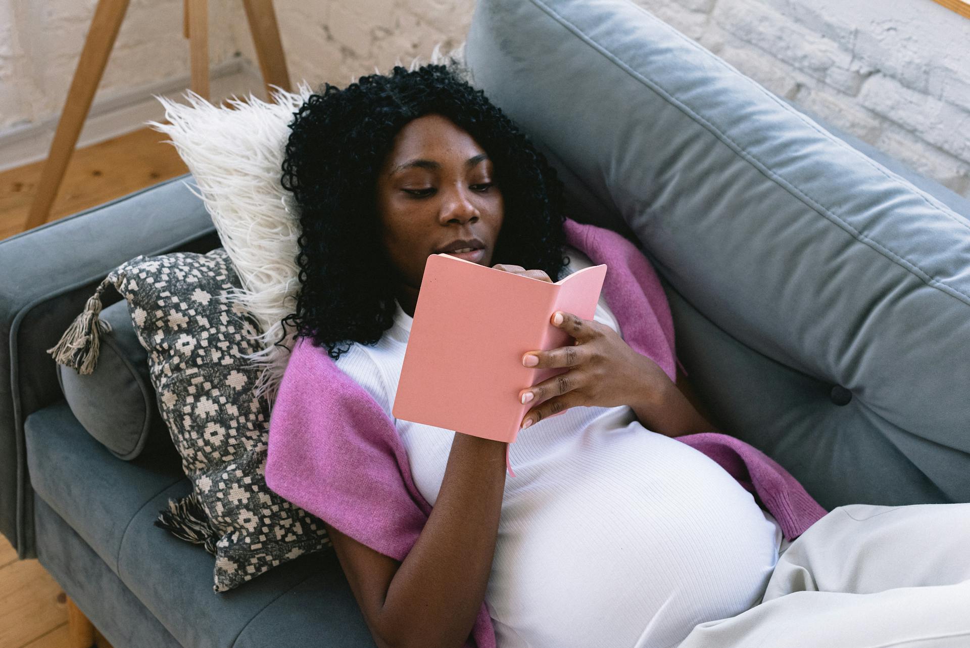 Bovenstaand beeld van een gefocuste zwangere Afro-Amerikaanse vrouw die aantekeningen maakt in een notitieblok terwijl ze rust op een comfortabele bank in de woonkamer