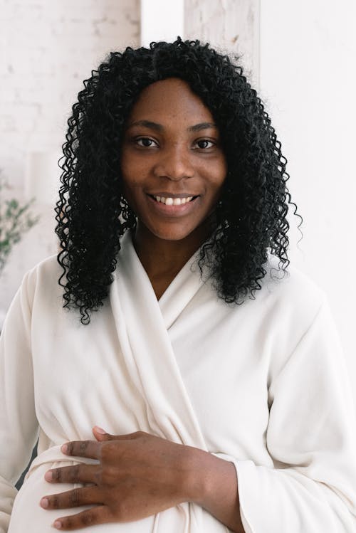 Free Cheerful pregnant African American female wearing bathrobe smiling and looking at camera while touching tummy in light room at home Stock Photo