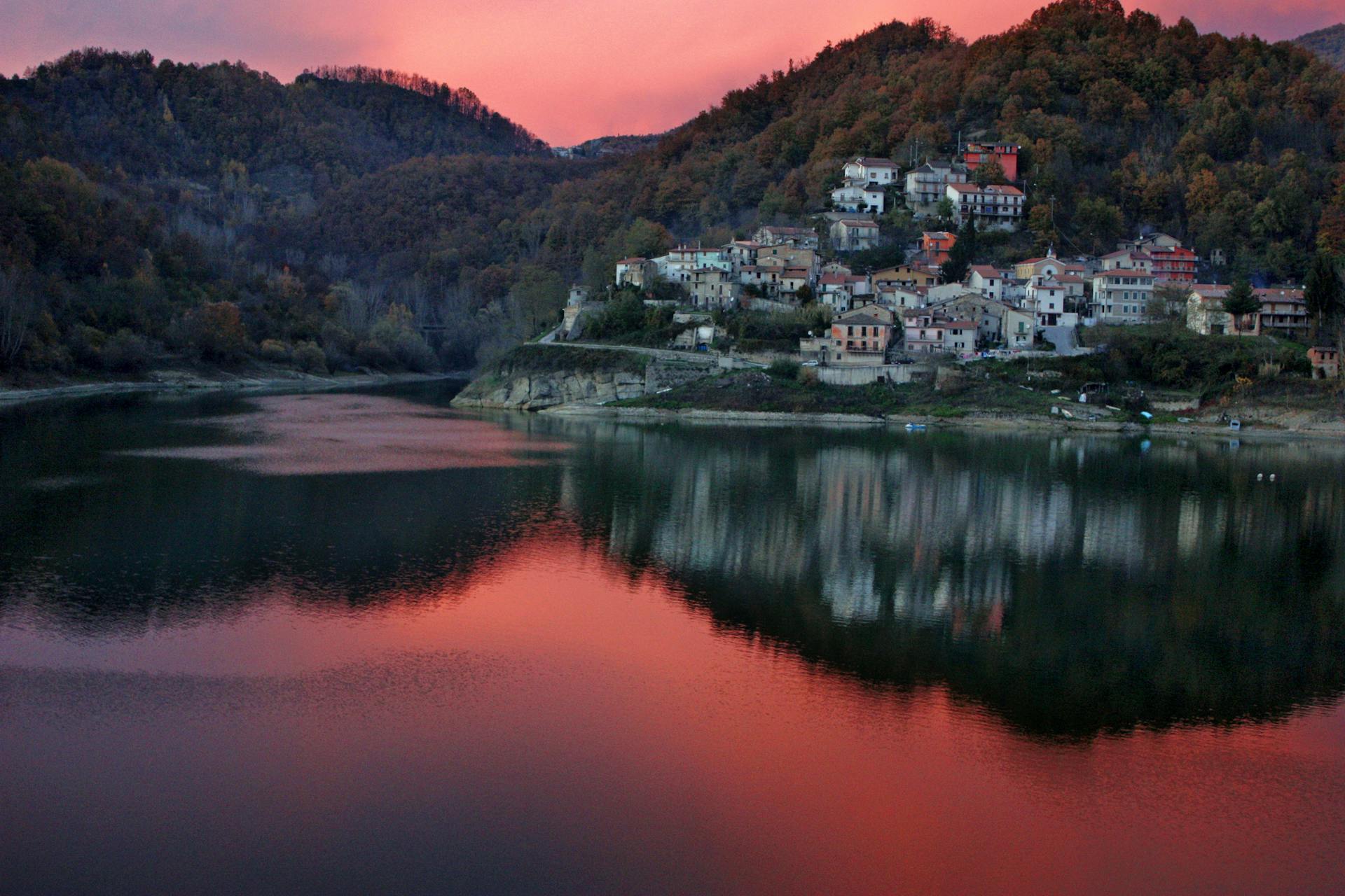 A charming village nestled in the mountains reflects in a tranquil lake at sunset, casting vibrant hues.