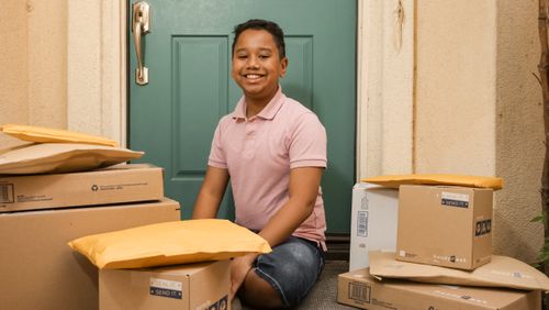 Free Young boy sitting on a porch surrounded by delivery packages, smiling warmly. Stock Photo