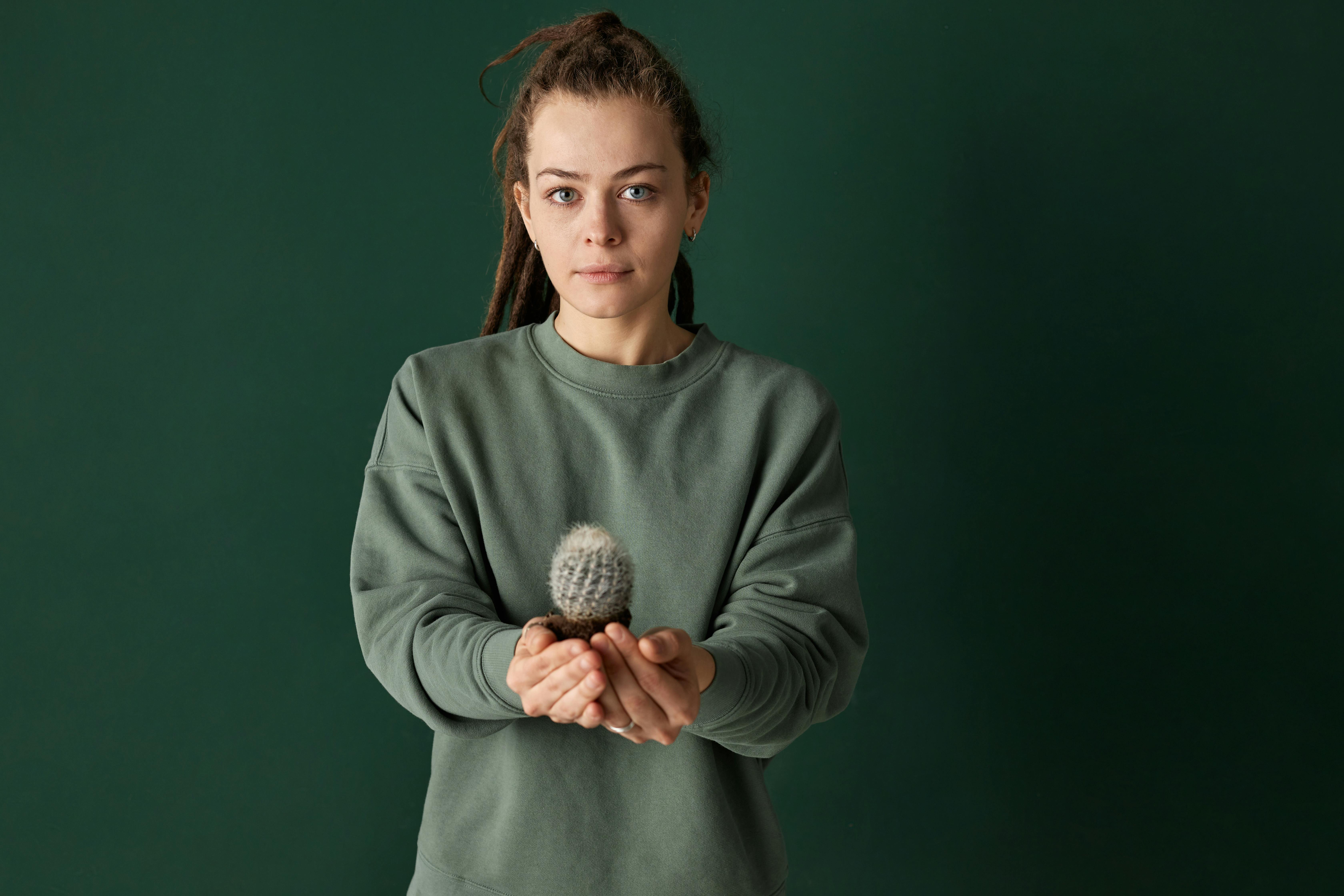 A young woman holding a small cactus, standing with a green background. Calm and natural mood.