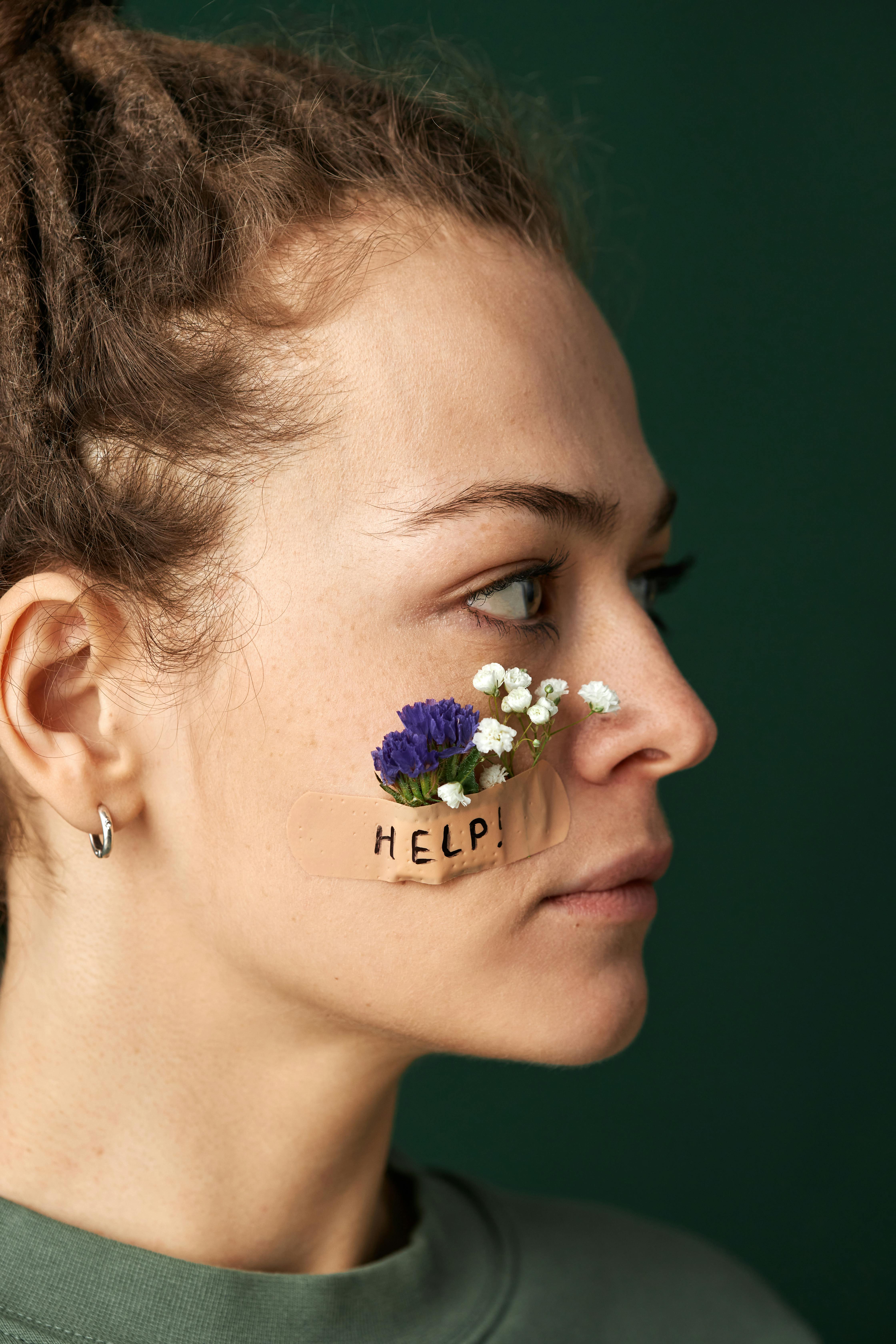 woman with flowers taped to her cheek and a sign help on a tape