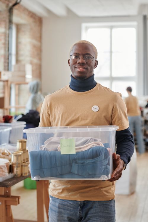 Free Black man volunteering in thrift store, holding donation box and smiling at camera. Stock Photo