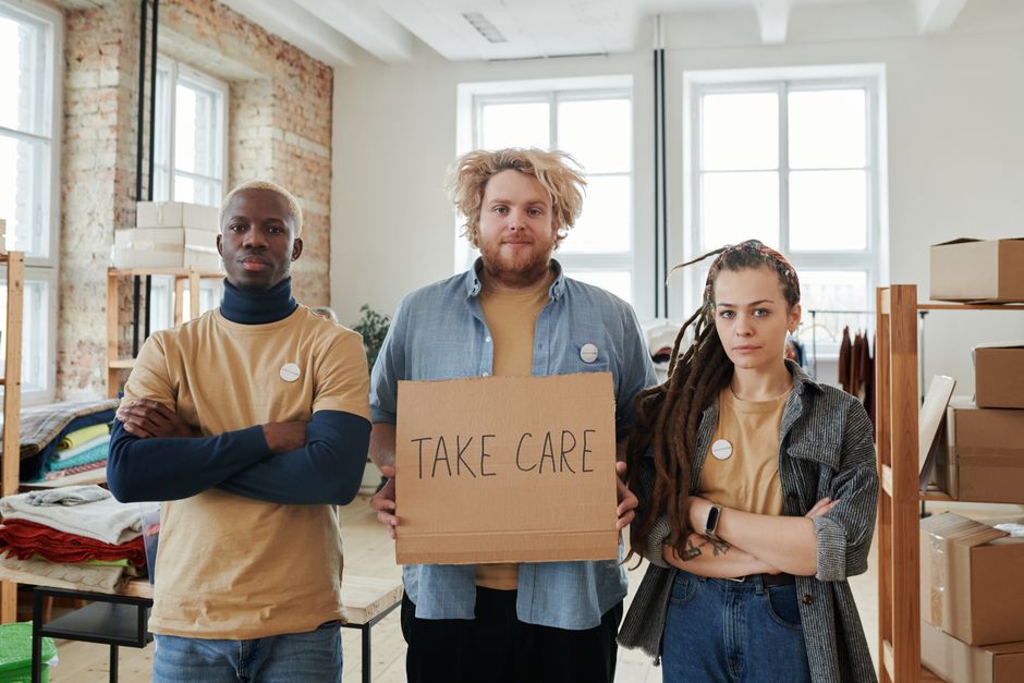 A diverse group of young adults holding a 'Take Care' sign inside a modern office.