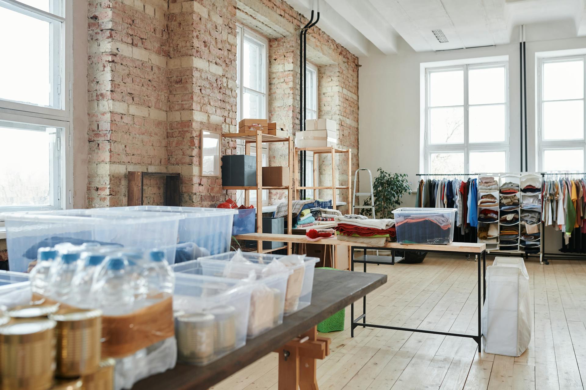 Spacious warehouse interior with wooden floors, racks of clothes, and storage boxes near large windows.