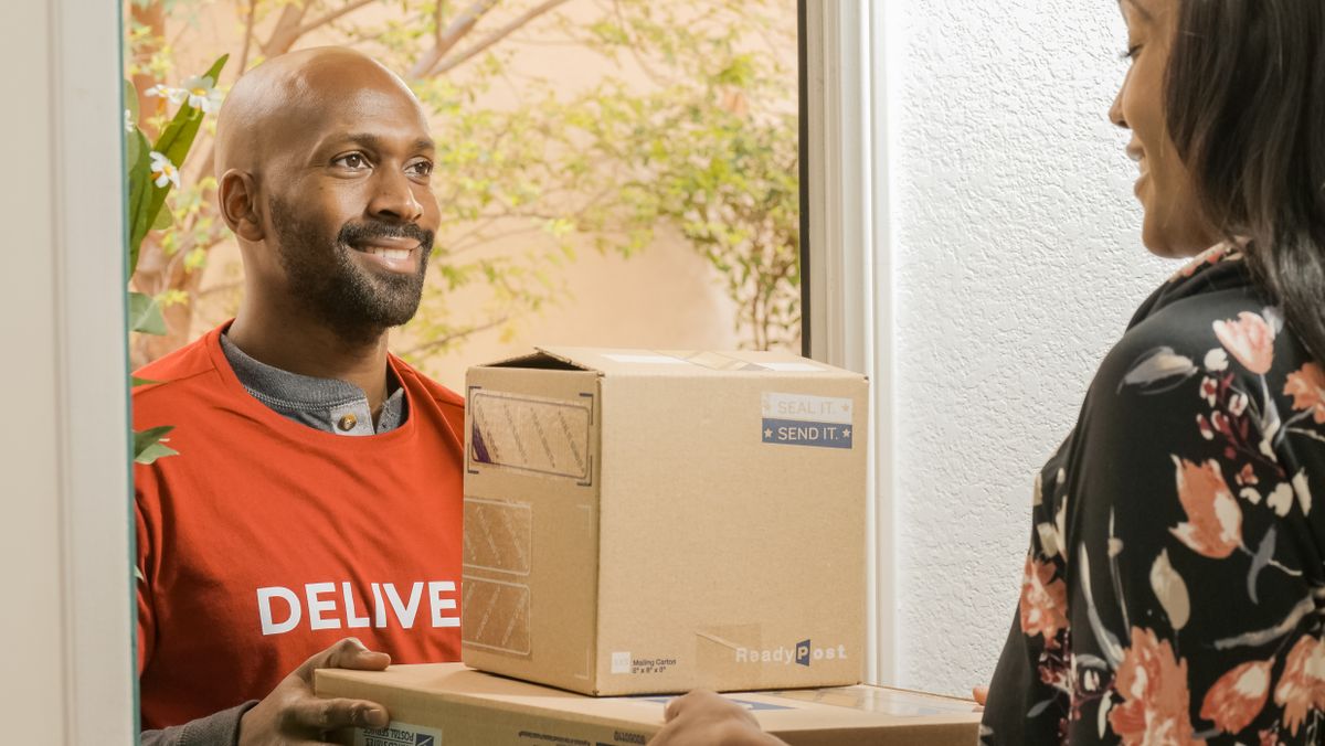 Entregador sorridente entregando uma caixa de encomenda para uma cliente na porta de casa