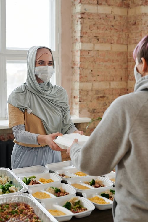 Free A woman wearing a hijab and face mask hands out packed food indoors, promoting charity and safety. Stock Photo