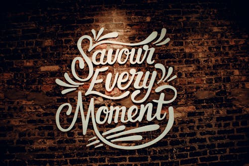 White creative signboard with Savour Every Moment inscription with decorative elements hanging on shabby brown brick wall of contemporary cafeteria