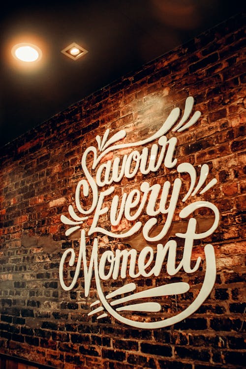 White signboard with Savour Every Moment inscription with creative design hanging on weathered brick wall in cafeteria with glowing lights