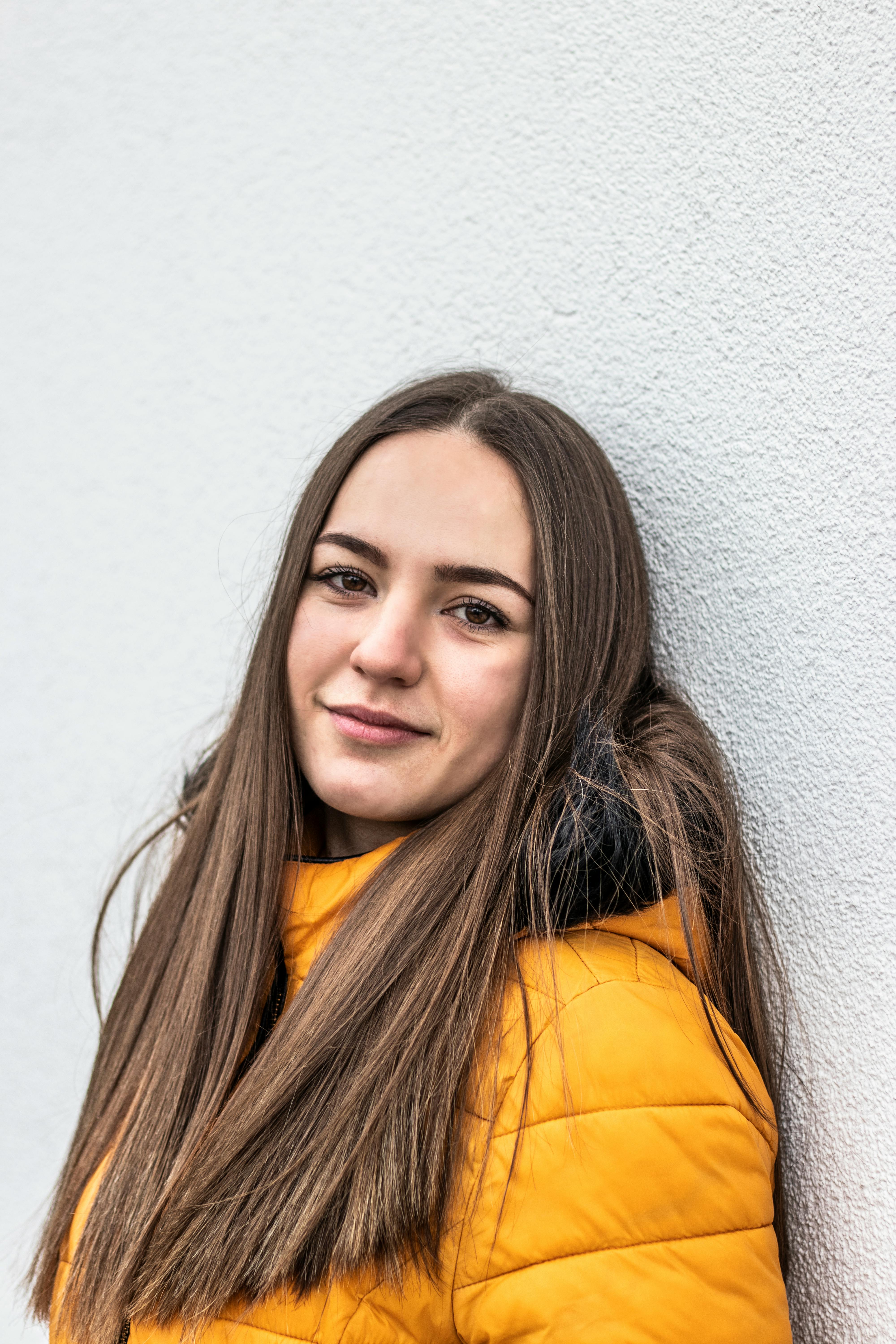 Portrait of a young woman with long hair in a bright winter coat against a white wall.