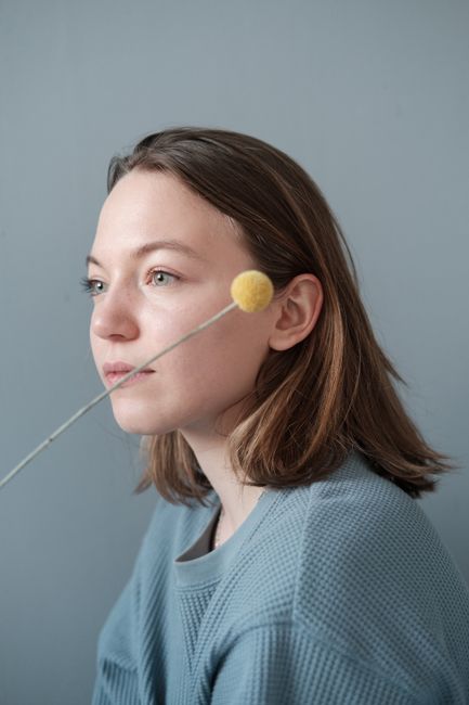 Young female in casual gray jumper with decorative yellow pompom on stick looking away thoughtfully while standing against gray background