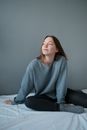Young beautiful woman in casual clothes sitting on unmade bed on white sheet in light room against gray wall and looking away