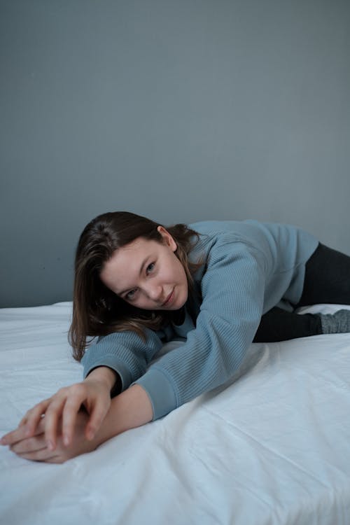 Young female in gray jumper lying on white bed sheet and looking at camera