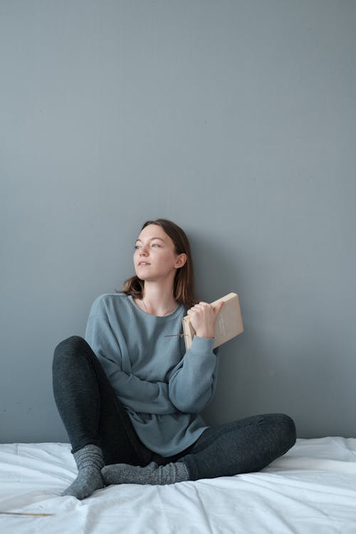 Full body of young female in gray clothes holding book bed and looking away while resting on white bed sheet in modern apartment