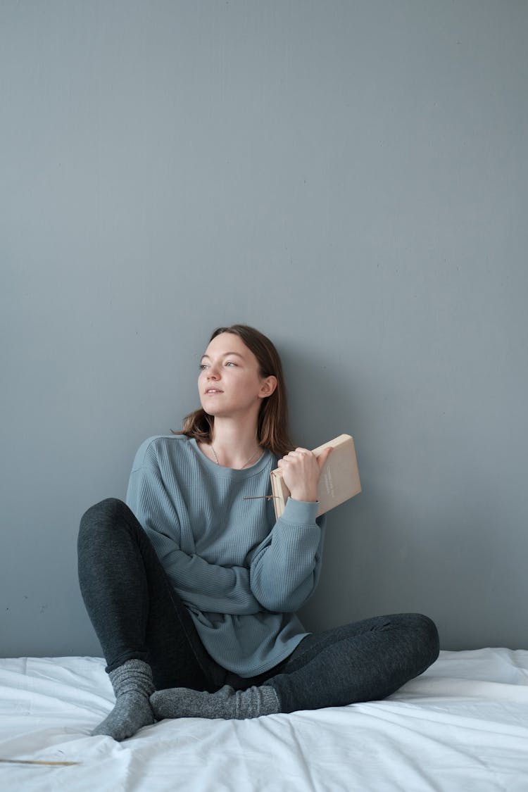 Young Woman In Warm Clothes Sitting On Unmade Bed And Holding Book