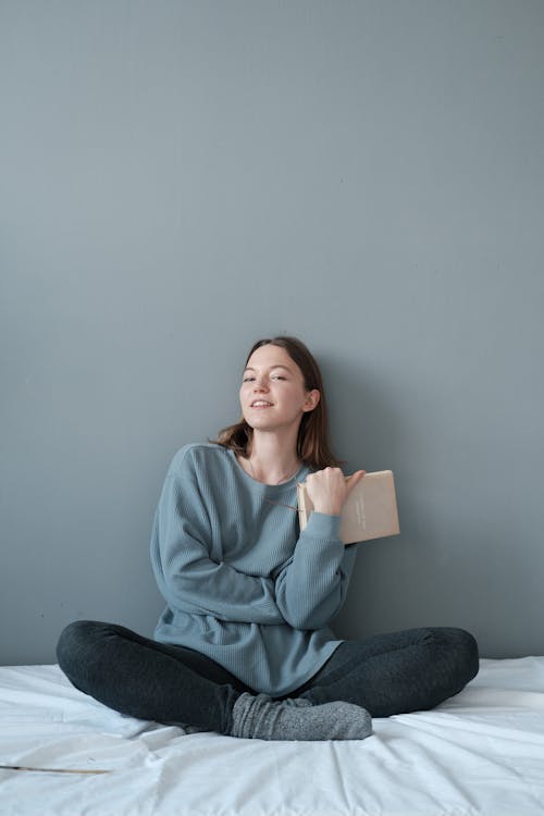 Young content female with textbook sitting with crossed legs on crumpled bed sheet while looking at camera on gray background